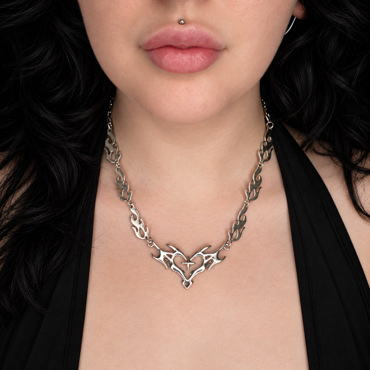 Model wearing the Heartburn necklace, featuring a silver-tone open heart pendant surrounded by symmetrical flame-like detailing on a matching flame-link chain.