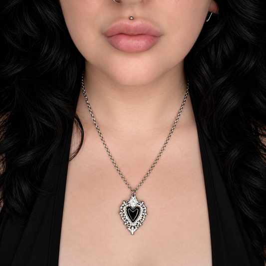 Model wearing the Memento necklace, featuring an ornate silver-tone heart-shaped pendant with a black heart centre and decorative detailing on a chain.