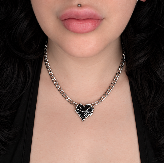 Model wearing the Fractured Heart necklace, featuring a glossy black broken heart pendant with silver-tone cracked detailing on a chunky silver-tone chain.